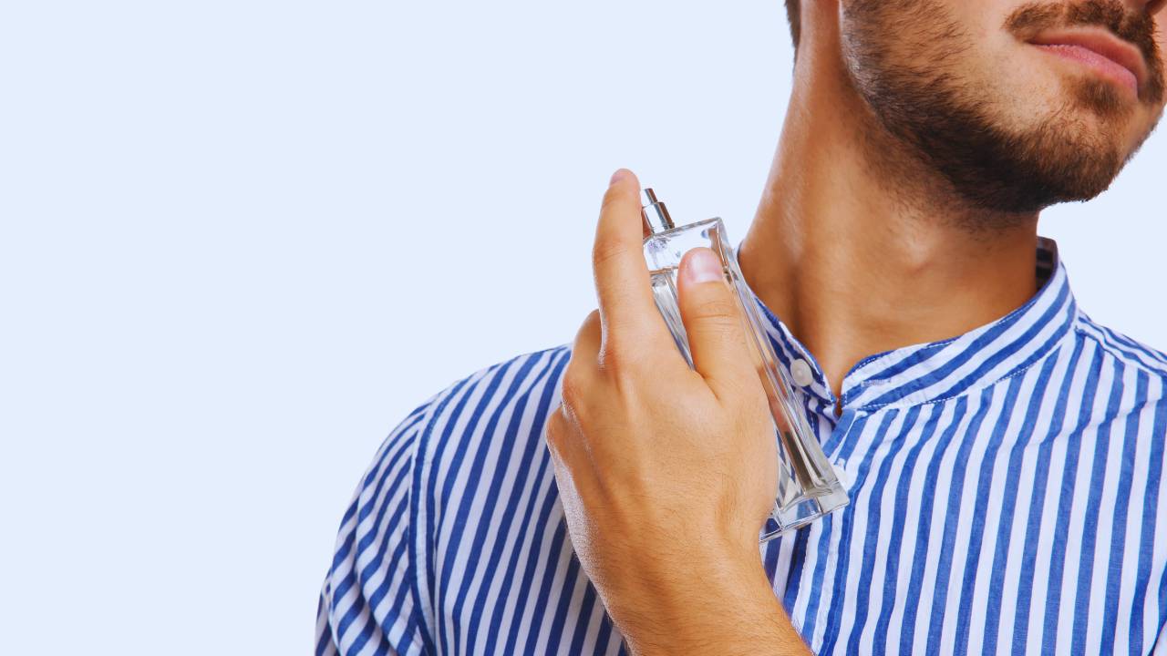 Homem de camisa azul e branca espirrando perfume no próprio pescoço