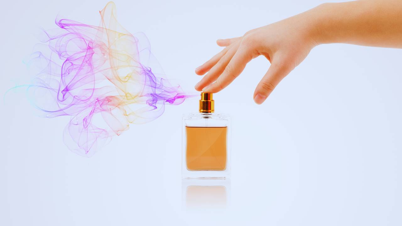 Mão feminina acionando o spray de um frasco de perfume, que solta uma nuvem aromática colorida