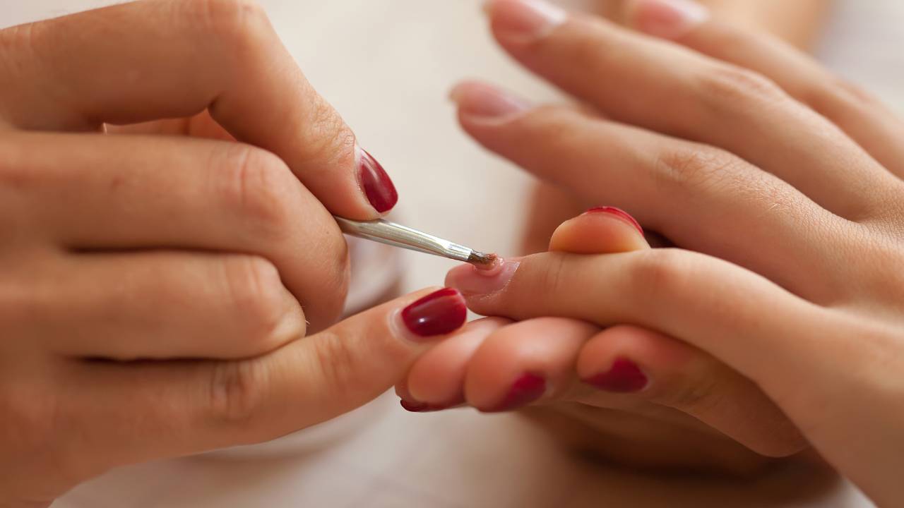 Um parte de mãos femininas segura e passa esmalte na mão de outra mulher