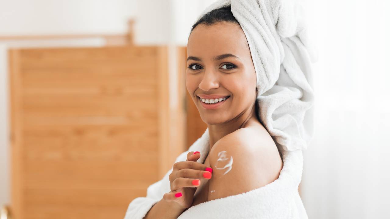 Mulher com toalha de banho enrolada na cabeça passando creme nos ombros