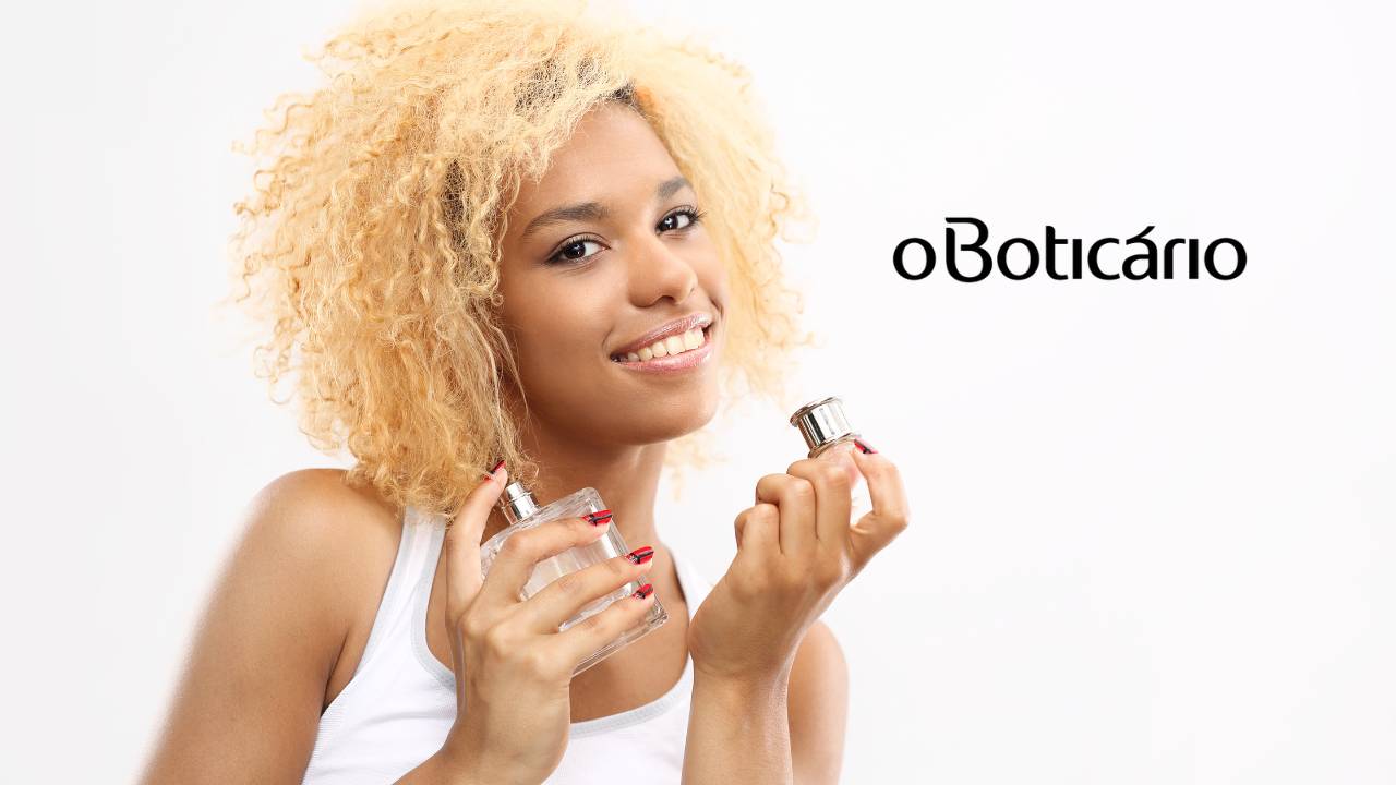 Mulher negra de cabelos loiros segura um frasco de perfume sorrindo; ao seu lado a logo do Boticário