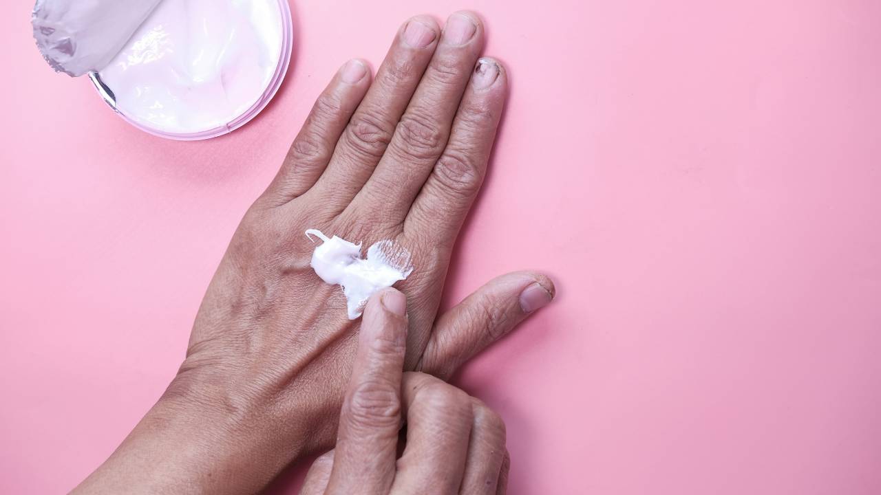 Mãos femininas sobre uma superfície rosa; a mão direita espalha creme nas costas da mão esquerda