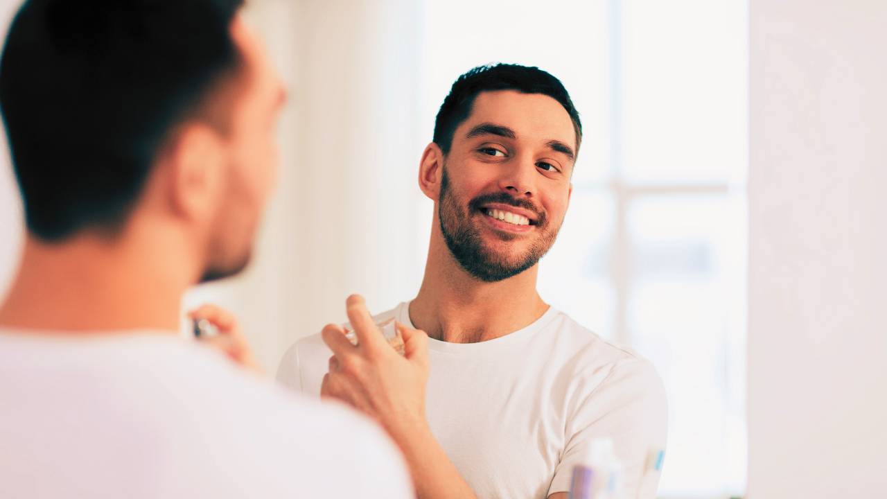 Homem sorridente passando perfume em frente ao espelho